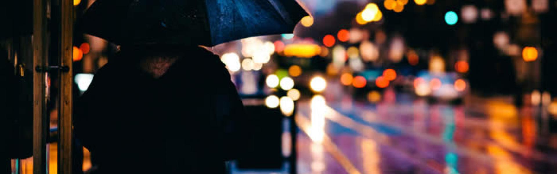 At dusk where in a rainy city scene where the are car taillights reflecting off of the wet surfaces of the road with a lone person standing near the camera with an umbrella. 